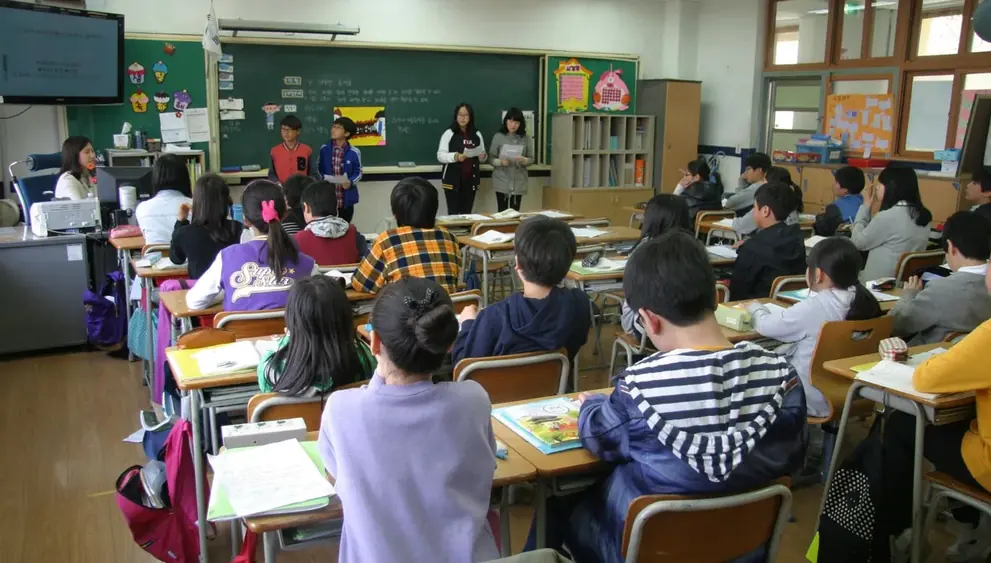 Sala de aula cheia vista do fundo, com alunos voltados para a lousa